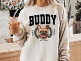 Person wearing a sweatshirt with a dog graphic and 'Buddy' text, holding a drink.